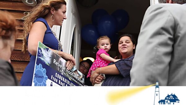A woman holding a small child stands in a doorway, smiling at another woman with a large sign celebrating the Revere Roof Rescue Program. Blue balloons and several people are in the background, with a lighthouse logo in the lower right corner.