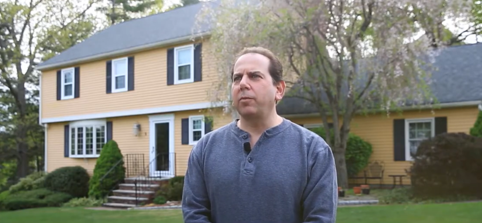 A man wearing a dark shirt stands outside in front of a large yellow house with white trim, black shutters, energy efficient windows, and a well-kept lawn with bushes and trees in Beverly.