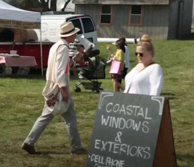 A man in a hat walks past a chalkboard sign reading Coastal Windows & Exteriors at The Vintage Bazaar outdoor market, while a woman in sunglasses strolls by. A red food truck and other people are visible in the background.