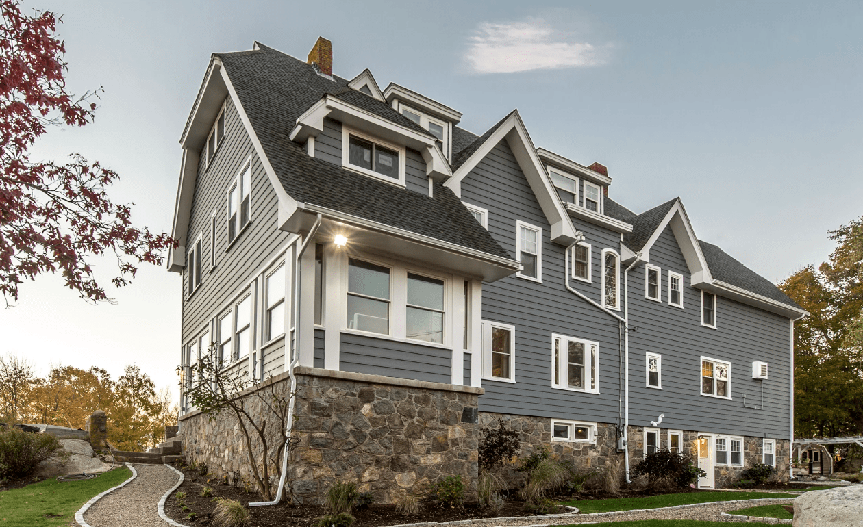 A large, three-story gray house with white trim and multiple windows showcases upgrades inspired by Cost vs Value 2024: Top 5 Upgrades to Maximize Your Homes ROI, featuring a stone foundation and manicured pathway under a clear sky with trees behind.
