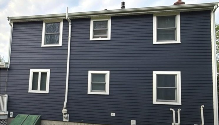 A two-story house with dark blue siding and white trim, featuring six windows and several exterior pipes on the back wall. The owner is HAPPY about his FREE ROOF. The sky is partly cloudy.
