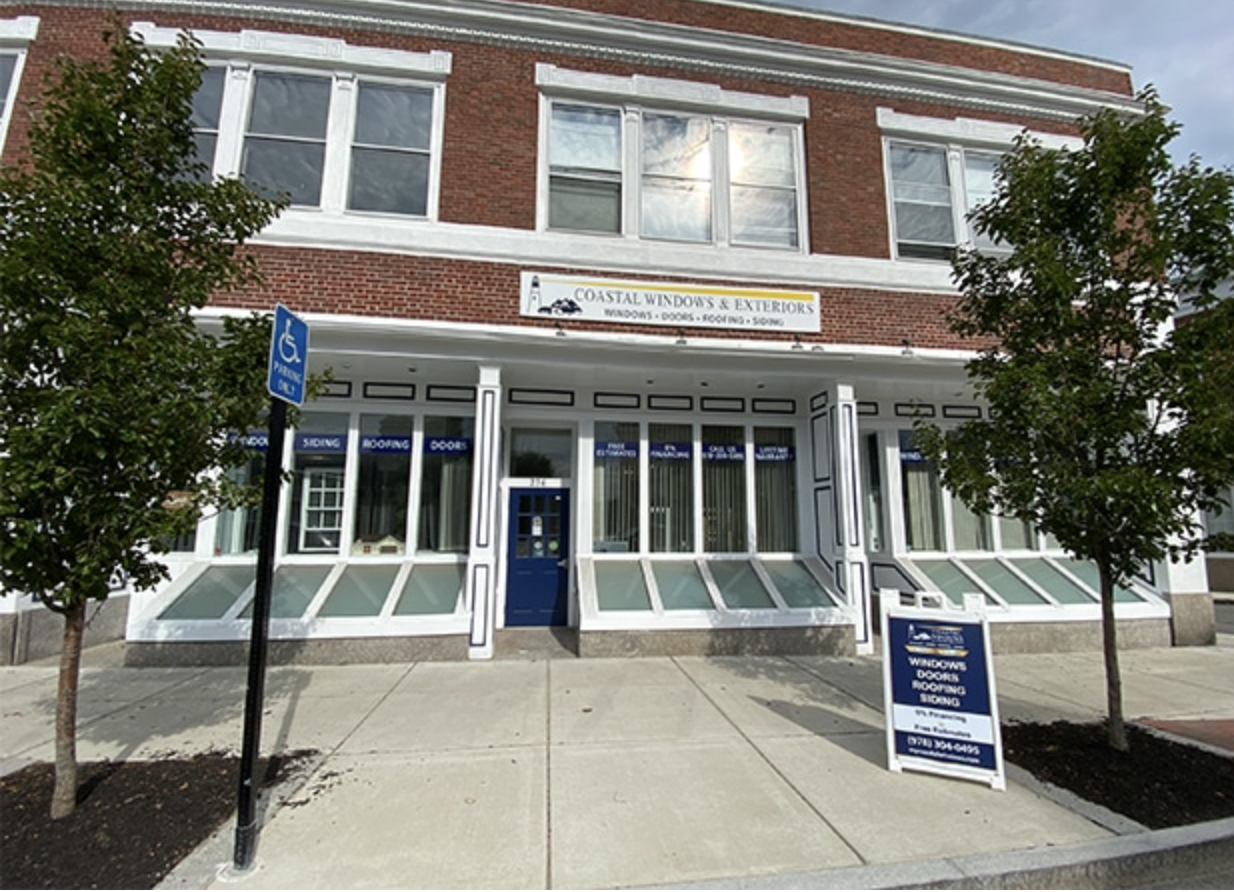 Street view of a brick building showcasing Coastal Windows & Exteriors with its inviting blue door and hanging sign. As spring home maintenance picks up, multiple windows catch the eye. Trees line the sidewalk, complemented by a blue handicap parking sign in front.