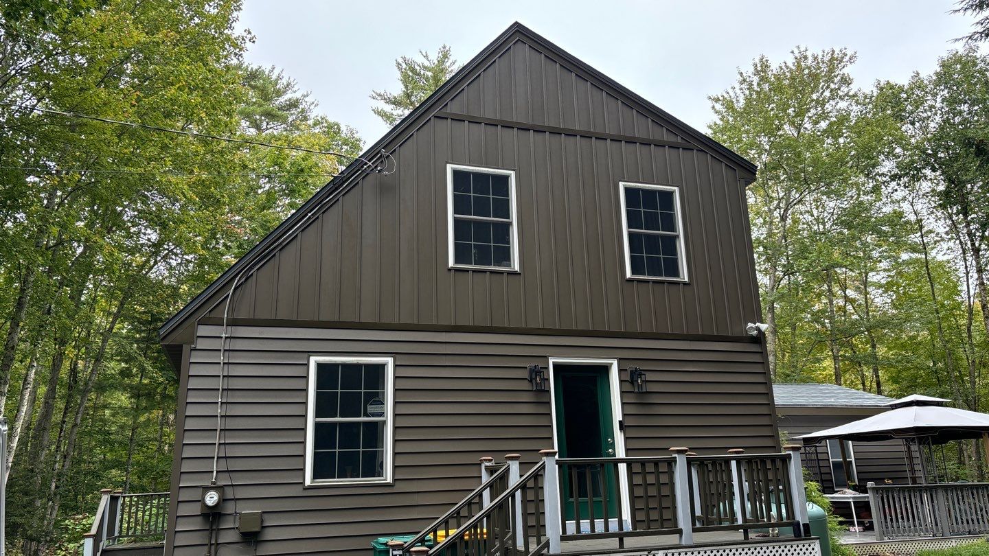 This two-story house, showcasing dark vinyl siding replacement in MA, boasts a steep gabled roof and is nestled among trees. It features two upper windows, a porch with railing, and a patio umbrella to the right. An overcast sky looms in the background.
