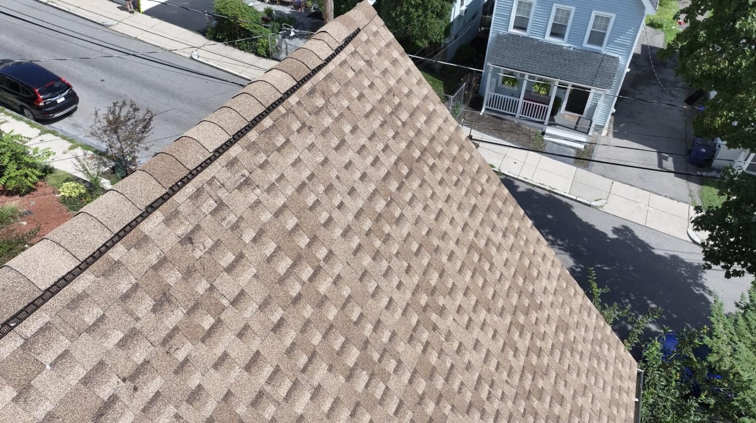 Aerial view of a house roof with tan shingles in a geometric pattern, perfect for your spring roof guide. Nearby, a street features a parked car and sidewalks beside a gray two-story house with a porch. Trees and vibrant greenery surround the area.