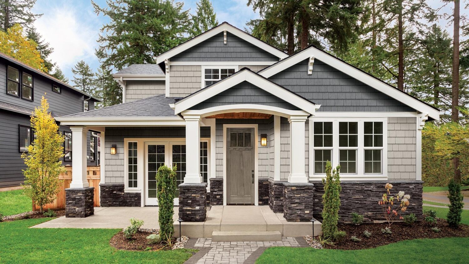Gray craftsman-style house with white trim and vinyl siding in Ipswitch, MA, featuring a covered front porch supported by white columns, gray front door, multiple windows, manicured shrubs, stone pathway, and tall trees in the background.