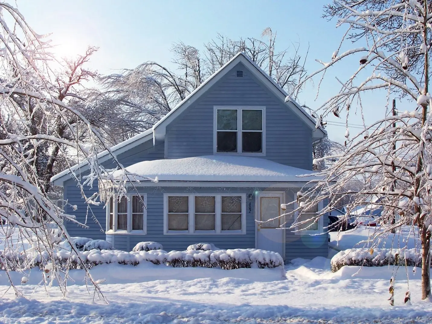 A blue house with snow-covered roof, yard, and trees sits under a clear blue sky on a sunny winter day. Icicles hang from the branches, and sunlight shines through the leafless trees.