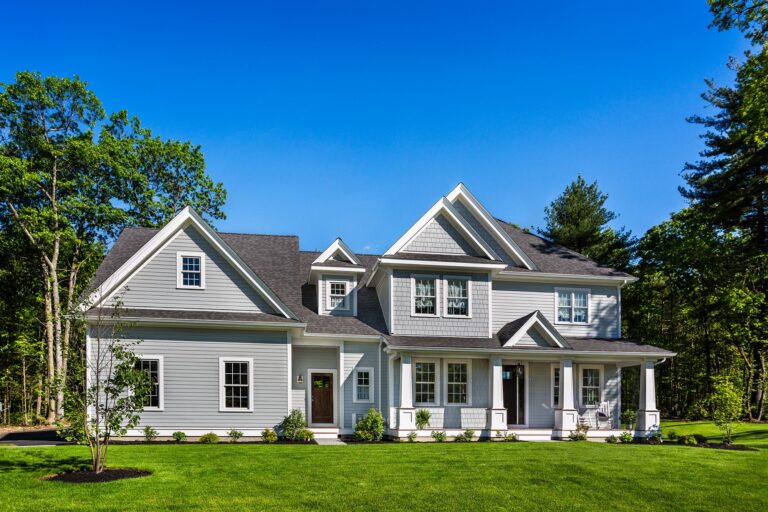 A large, two-story white house with gray trim and premium vinyl siding in Essex MA, featuring gabled roofs and a front porch, surrounded by green lawn and trees under a clear blue sky.