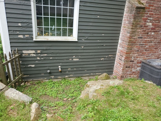Peeling paint and bare patches cover the lower wall of green wood siding beside a brick chimney and air conditioning unit. Our ultimate fall home maintenance checklist explains how damage like this lets moisture into the walls.