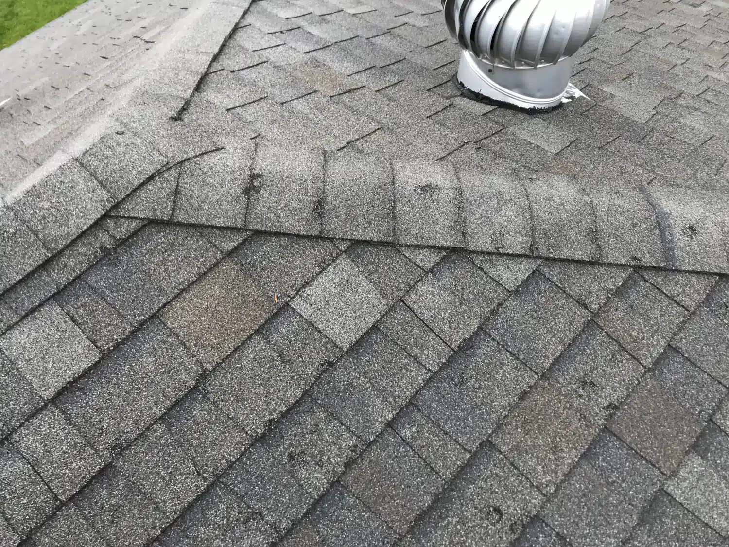 Dark streaks and dirty patches on gray asphalt shingles near a roof vent on a Wellesley, Greater Boston home.