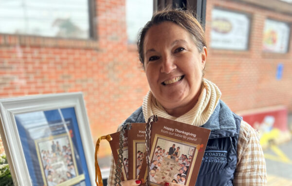 A smiling woman stands indoors by a window, holding several Thanksgiving-themed booklets. Showcasing “Freedom from Want” with Turkey Donation, she wears a checked shirt with a navy vest beside a display of framed pictures and colorful flowers.