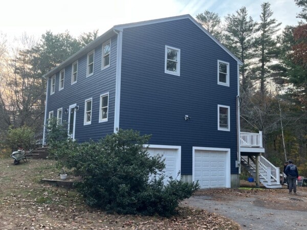 A two-story blue house with white trim features James Hardie Siding in Groveland, double garage doors, and a side staircase. Trees surround the home, and a person is visible near the driveway.