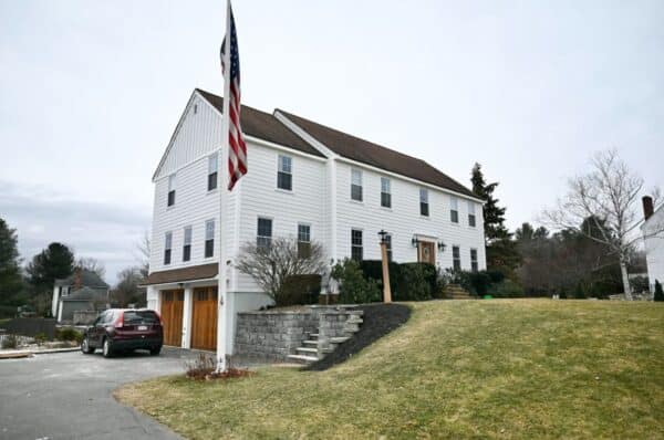 This two-story white house in North Andover features a brown roof, Colonial Makeover Siding, attached garage, raised front yard, and an American flag by the driveway. A car is parked nearby with trees in the background under an overcast sky.