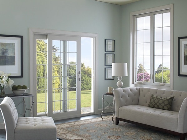 A cozy living room with light green walls, featuring a sofa and chair in gray tones. Large windows overlook a sunny garden. A white lamp sits on a side table, and framed artworks adorn the walls. A patterned rug covers the floor.