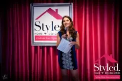 A woman in a blue dress holds a microphone and papers while speaking on stage at a Style Home & Design event supporting the Ellie Fund. She stands in front of a red curtain and a sign that reads Style Home & Design.