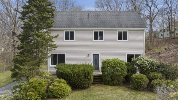 This charming two-story house in Ashland, MA, features beige vinyl siding surrounded by lush green shrubs and trees. It boasts a dark, steeply pitched roof and five visible windows. A narrow pathway leads to the inviting front door, all set against a serene wooded backdrop.