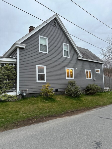 A gray house with white trim, featuring James Hardie siding, boasts a steep roof and sits alongside a road in Essex, MA. The windows are warmly lit from inside. Small shrubs and a grassy lawn embellish the front, with overcast skies looming above.