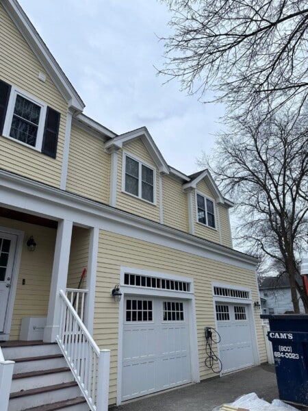 A yellow house with white trim stands proudly in Needham, MA. It boasts two stories and features three dormer windows above two garage doors. A set of stairs with white railings welcomes you to the front door. To the right, a blue dumpster hints at recent James Hardie Installation work amid leafless trees.