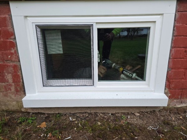 A small rectangular basement window with a white frame is set in a red brick wall. The window, possibly a replacement window from North Attleboro, has two panes; one is screened. A person is holding a caulking gun near the window from the outside. A grassy area is visible through the glass.