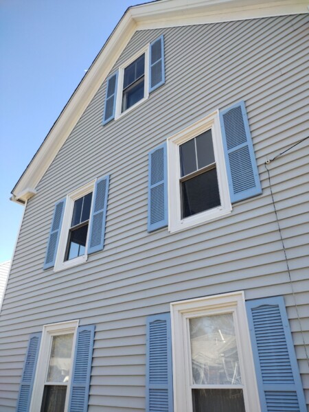 A charming house in Kennebunk features light blue horizontal siding and crisp white trim, punctuated by windows with matching blue shutters. The sky is clear and bright, enhancing the serene vibe of this delightful home.