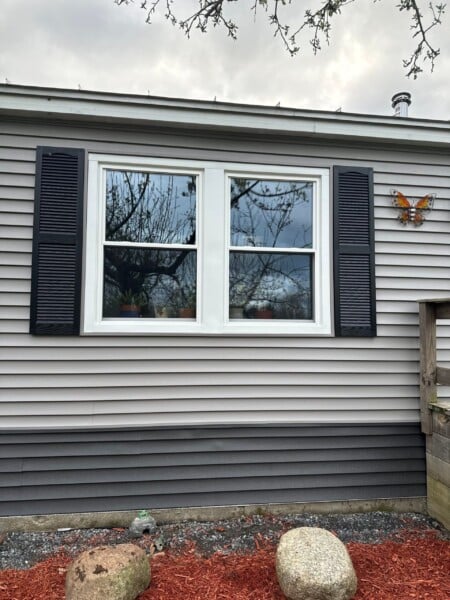 A house in Winslow, ME, boasts elegant beige siding and replacement windows with striking black shutters. A decorative butterfly hangs on the wall beside the window, overlooking a small garden adorned with mulch, two large rocks, and a designed stone.