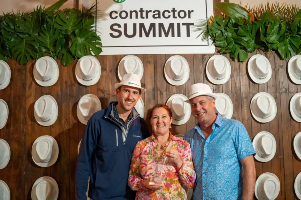 Three people stand in front of a wooden wall, wearing white hats, with a sign reading Contractor Summit. They are smiling amidst a tropical backdrop, embodying the spirit of service excellence that defines the James Hardie ethos.