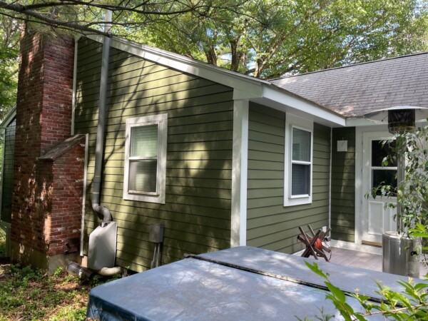 A charming small green house with white trim, nestled among trees in Andover, MA. It features a brick chimney and siding with two windows on the side. A hot tub with a cover sits on the deck leading to a white door, perfect for your next home improvement project.