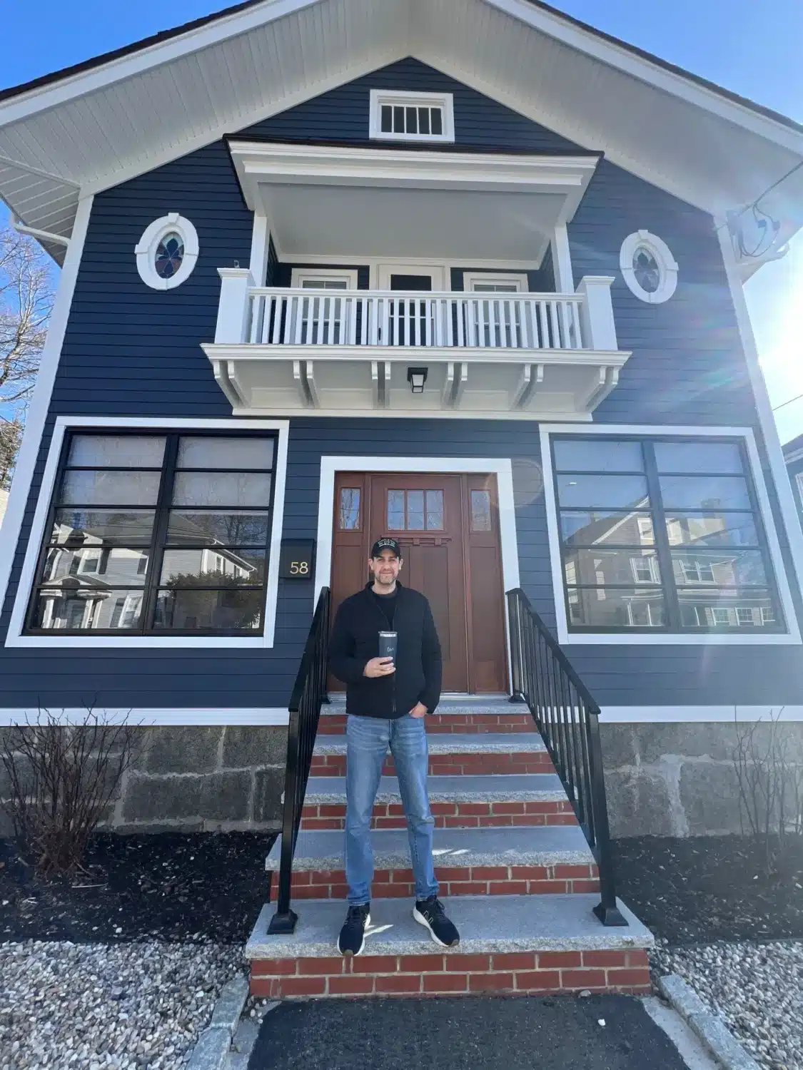A person stands on the steps of a two-story Ocean Blue house with white trim, holding a drink. The clapboard siding complements the large windows, wooden front door, and small balcony above the entrance. Its a clear day with no visible clouds.