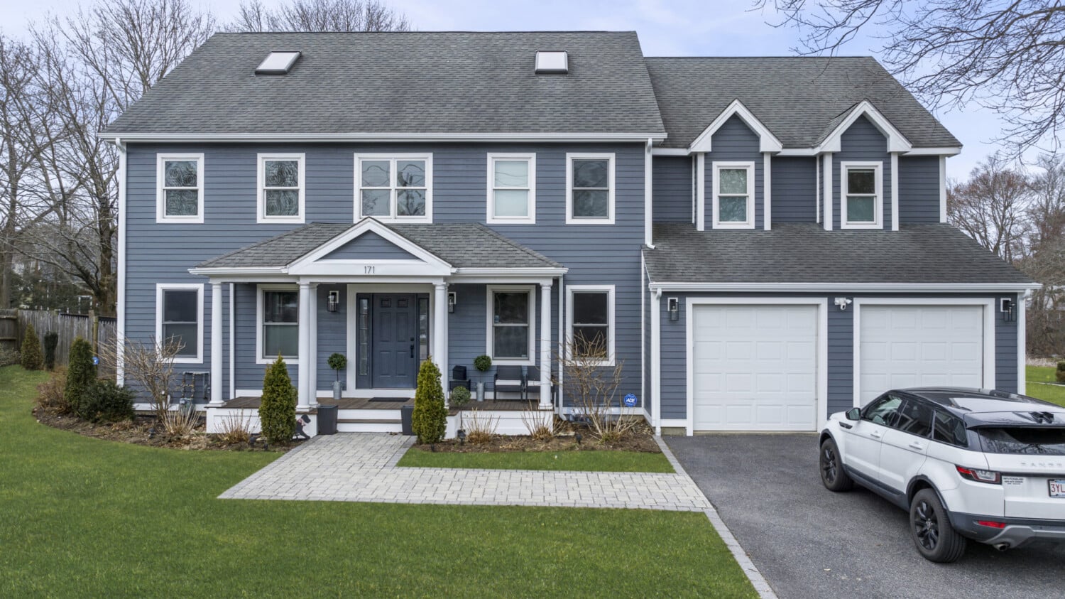 A two-story blue house with white trim and Pearl Gray Cedarmill Siding features multiple windows and a double garage. A white vehicle is parked in the driveway, and the front lawn is neatly maintained. The sky is clear, showcasing trees in the background with a touch of James Hardie elegance.