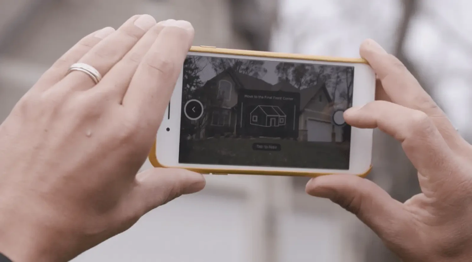 A person is holding a smartphone with a yellow case, capturing or viewing a house on the screen. Overlay controls suggest its a virtual appointment experience, ensuring no contact while exploring the property.