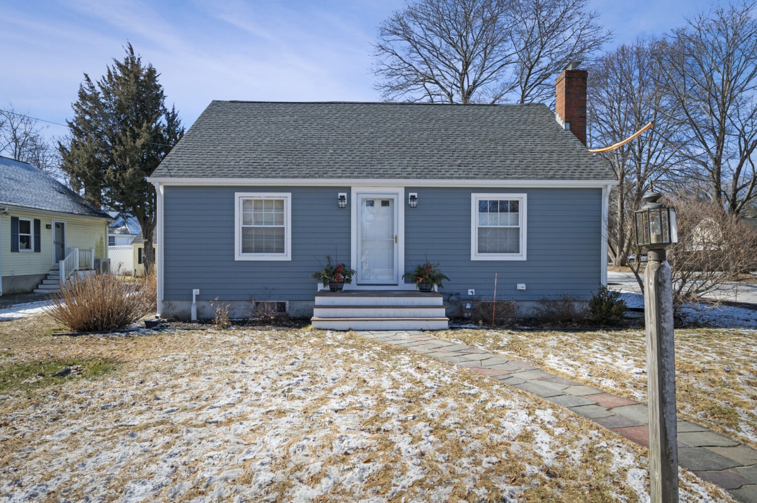A small, Boothbay Blue single-story house with white trim and a dark gray roof. Featuring a centered front door with steps and two vinyl window replacements. The yard is partly covered in snow patches, and a stone pathway leads to the entrance, framed by bare trees in the background.