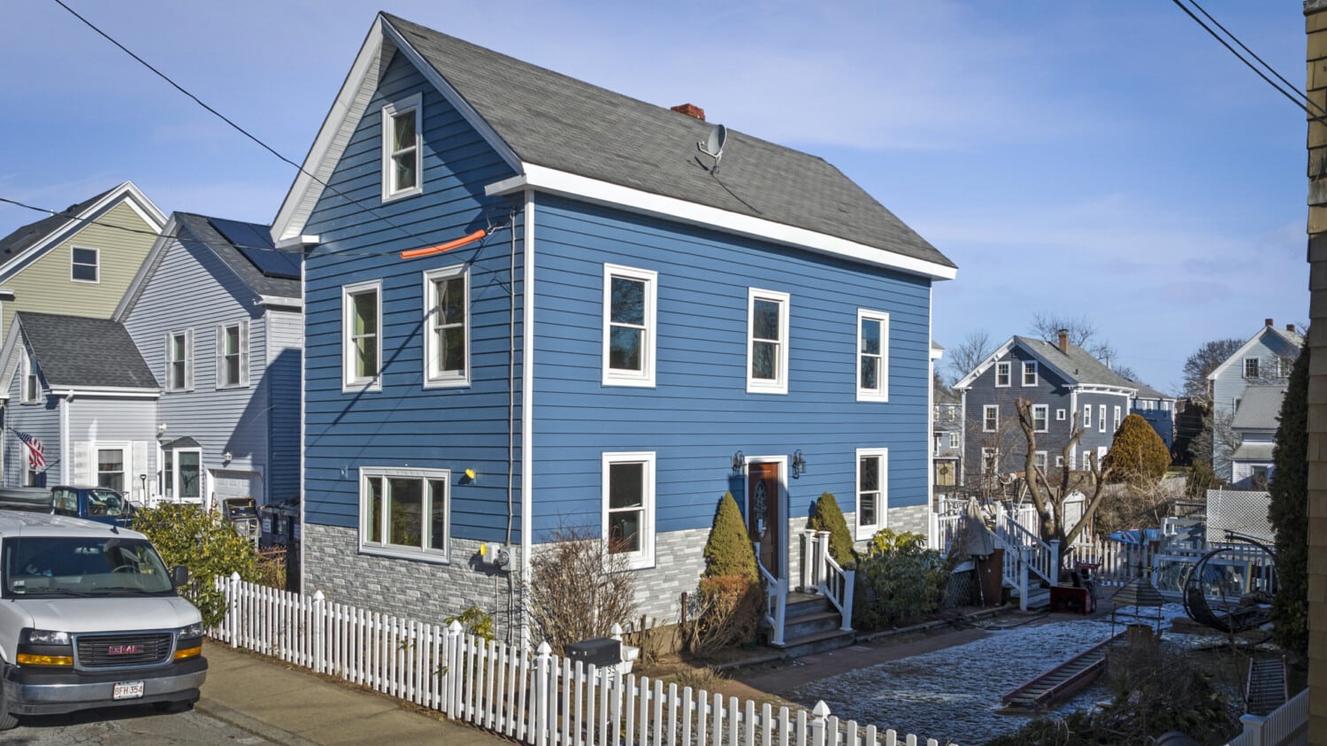 A two-story blue house with vinyl siding and a fenced yard graces a suburban street in Salem, MA. Featuring white trim, several windows, and a small porch with steps, it stands charmingly surrounded by its white picket fence. Nearby, a grey SUV is parked on the road.
