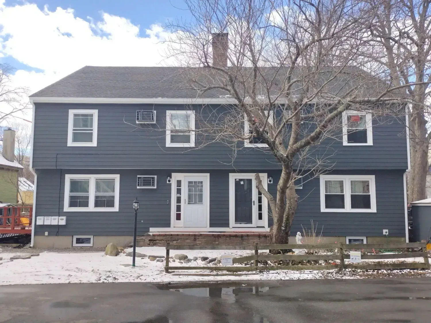 A two-story gray house with white trim, expertly updated by James Hardie® Siding Contractors in Salem MA, features two front doors and multiple windows. A leafless tree stands in the partly snowy yard, bordered by a wooden fence along the driveway under a partly cloudy sky.