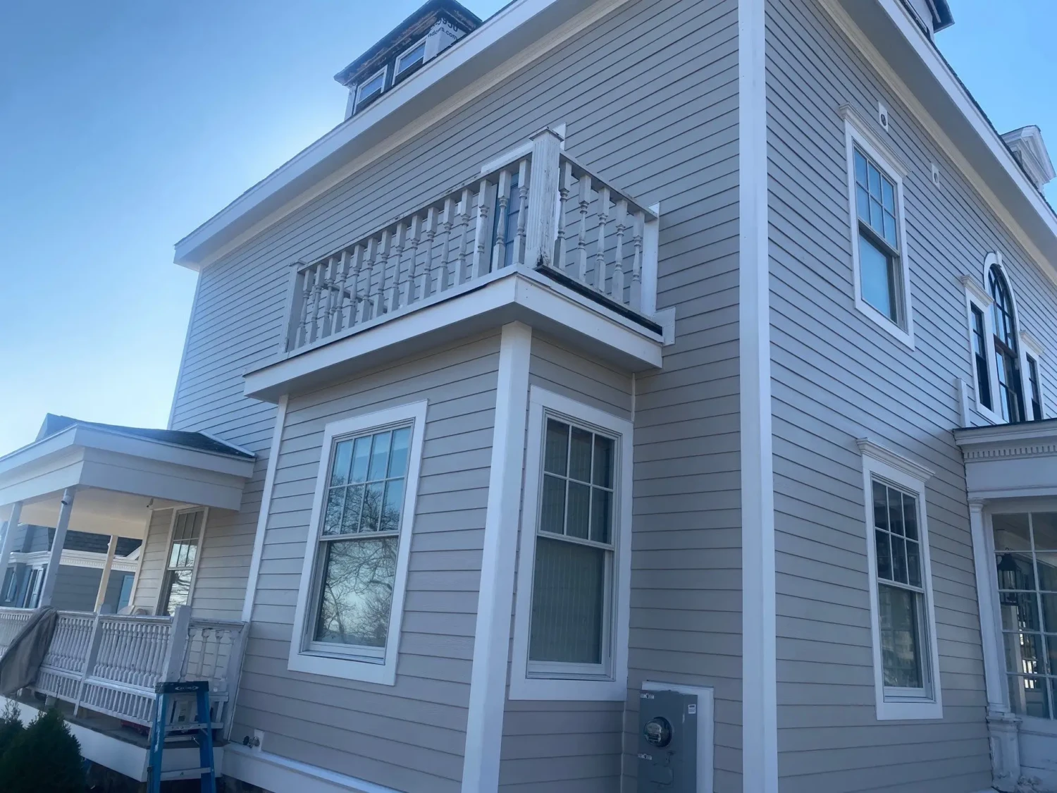 A two-story beige house in Beverly, MA with white trim and a small balcony boasting decorative railings on the second floor. Several windows grace each level, and a ladder hints at recent work by local contractors, perhaps installing James Hardie siding. The sky above is clear and blue.