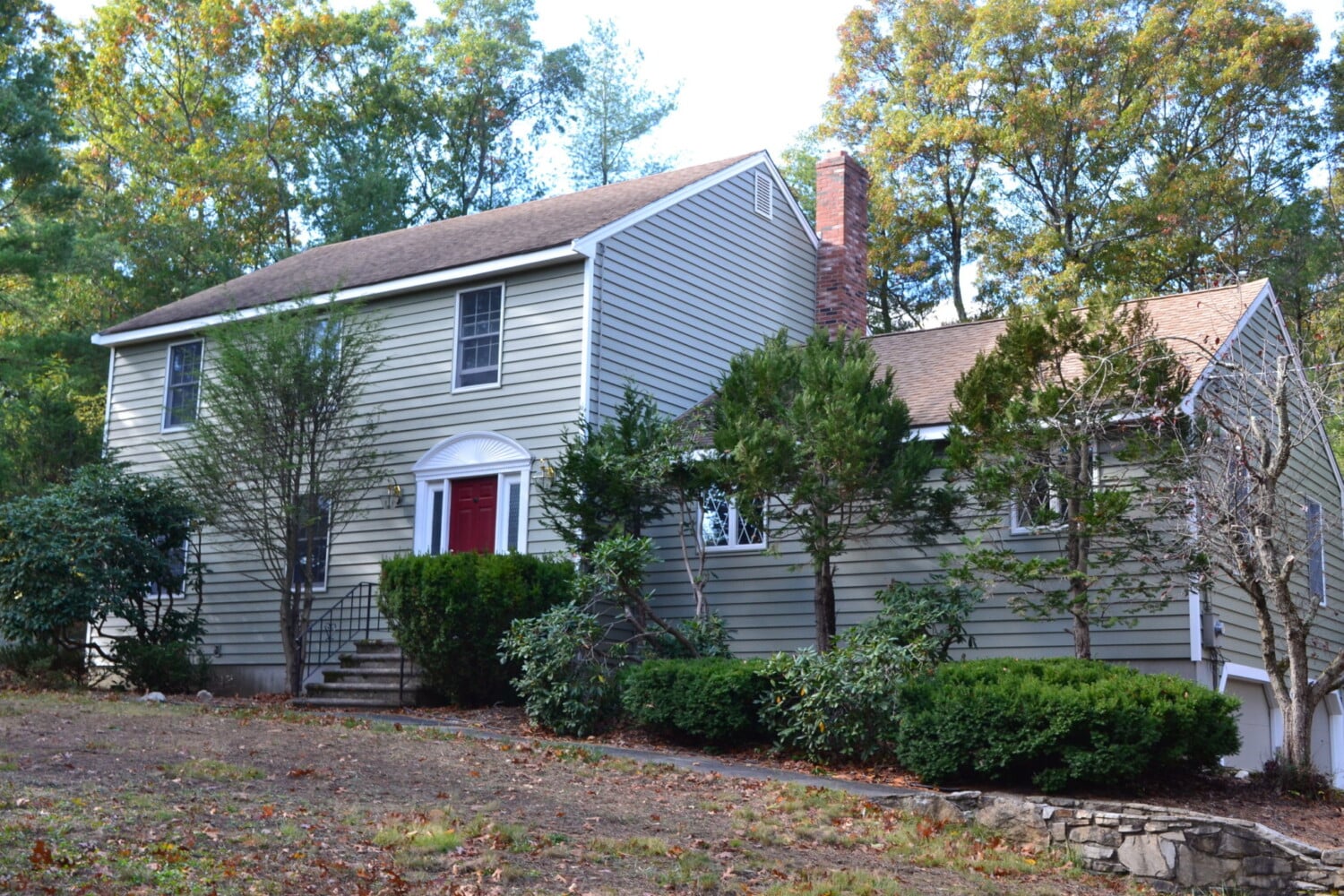 A charming two-story house in Franklin, MA, features light gray siding and a vibrant red front door. Surrounded by lush trees and shrubs, it boasts a brown roof with a brick chimney. The front yard enjoys a refresh with its sloped grassy area framed by a classic stone retaining wall.