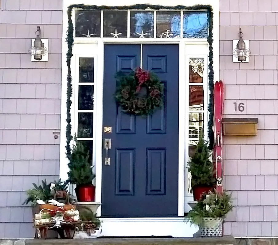 A festive entryway featured in Boston Magazine showcases a navy blue front door adorned with a holiday wreath. Small evergreen trees in red pots and red skis enhance the entrance, while garland and star-shaped lights frame it. A sled leans against the left side, completing the charming scene.