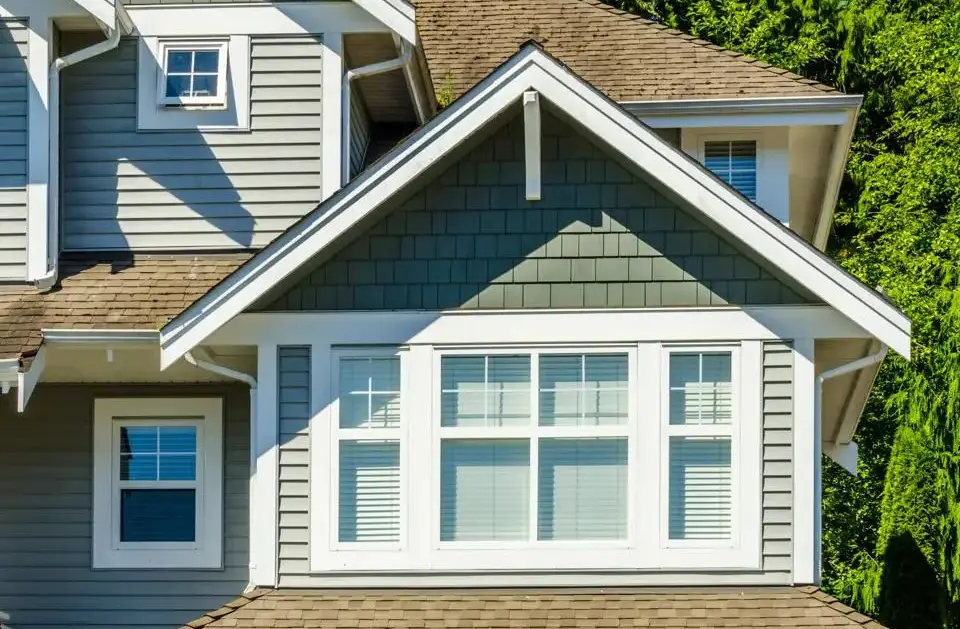 Close-up of a house exterior with gray siding and white trim. A gabled roof with a small window and a larger, multi-pane window that might soon need to replace old windows is visible. The background features lush green trees partially in sunlight, reminiscent of scenes from the Boston Globe.