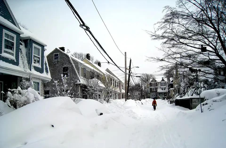 A person wearing an orange jacket walks down a snow-covered residential street lined with houses and leafless trees, capturing a quintessential winter scene. Snowbanks are piled high, and the sky is overcast, reminiscent of a Boston Magazine cover. Power lines stretch across the serene landscape.