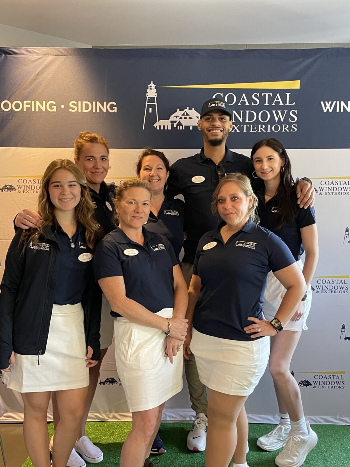 A group of seven smiling employees in navy shirts and white bottoms stands in front of a Coastal Windows & Exteriors banner at an indoor WBL PGA Golf Event.