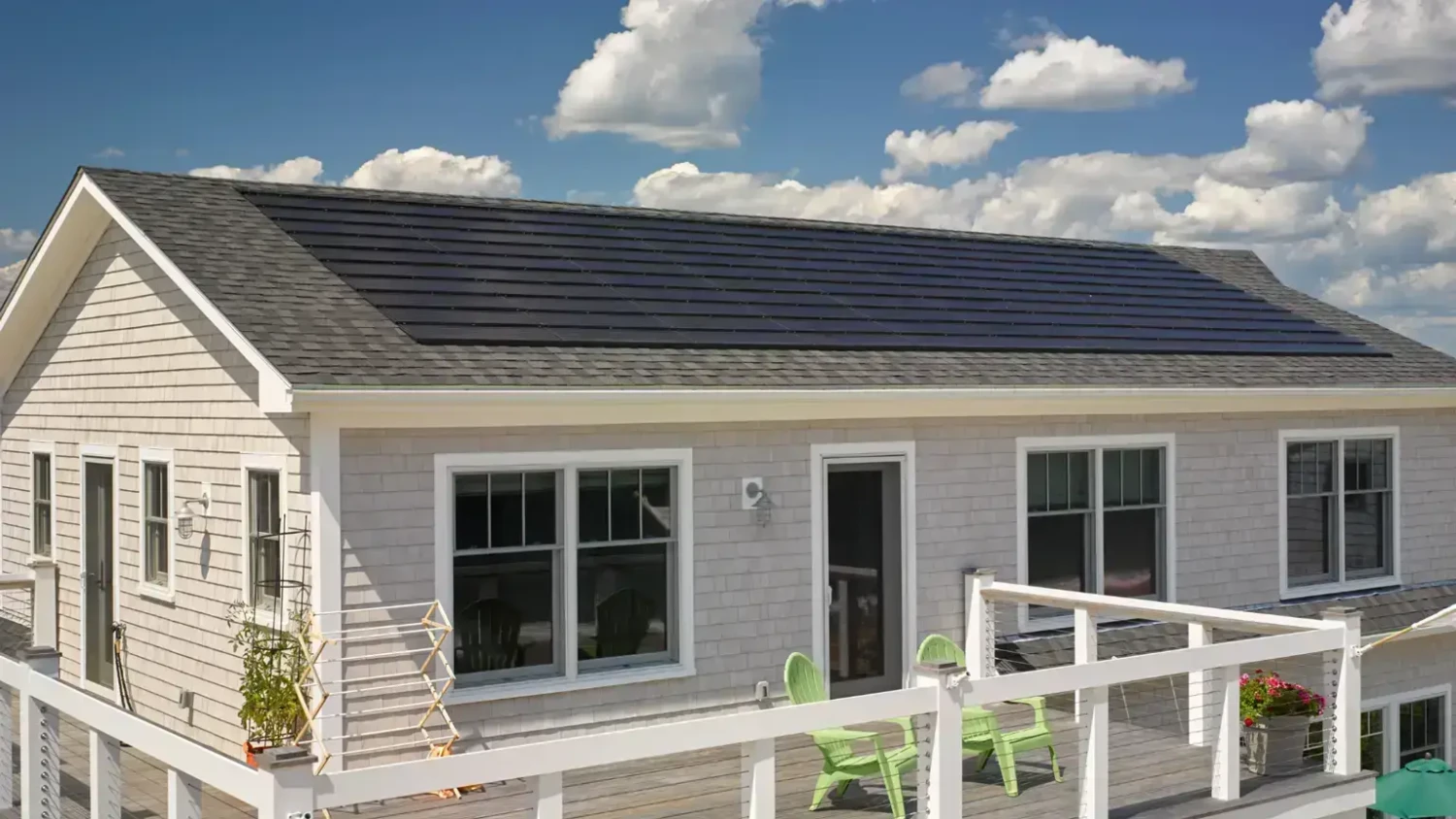 A small house features a sloped roof with solar panels. The exterior is light-colored with two windows and a central door. Green chairs and a table are on the wooden deck with a white railing. Fluffy clouds fill the blue sky above.