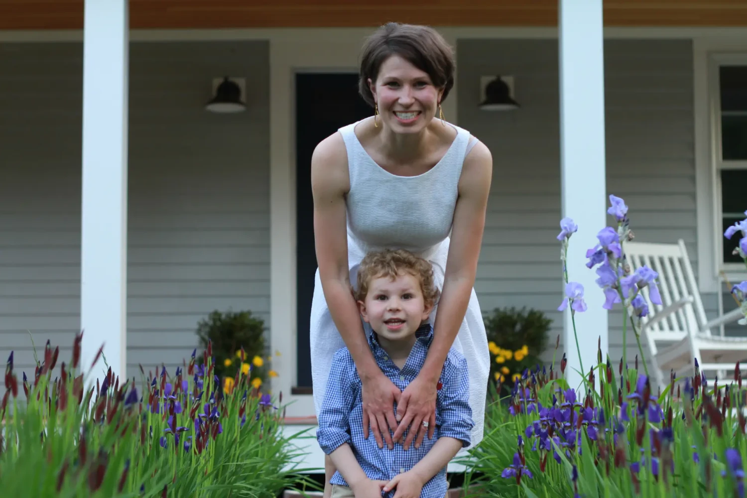 A woman in a light dress bends down and smiles behind a young boy with curly hair. They are outdoors at Latitude 42, in a garden with purple flowers and a house with a porch in the background.