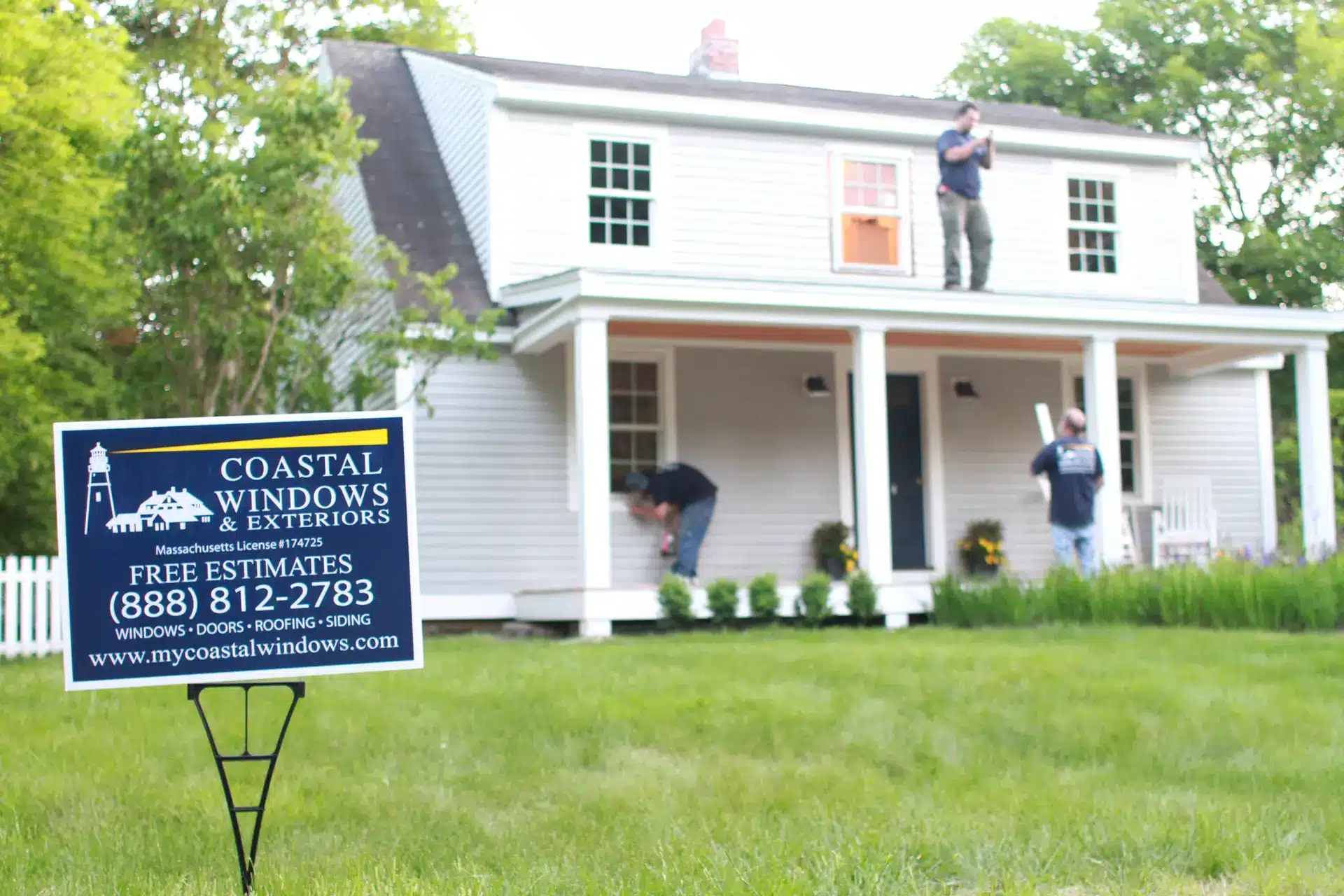 Three workers are installing windows on a white two-story house in this New Construction Window Time Lapse. A sign on the lawn reads “Coastal Windows & Exteriors: Free Estimates,” with contact info and a website amid lush green grass and trees.