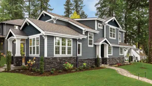 A modern two-story house with dark gray siding and white trim, featuring large windows. The lower part of the facade has a stone exterior. Its surrounded by a well-kept lawn, trees, and a paved walkway leading to the entrance—ideal for any New England homeowners siding inspection checklist.