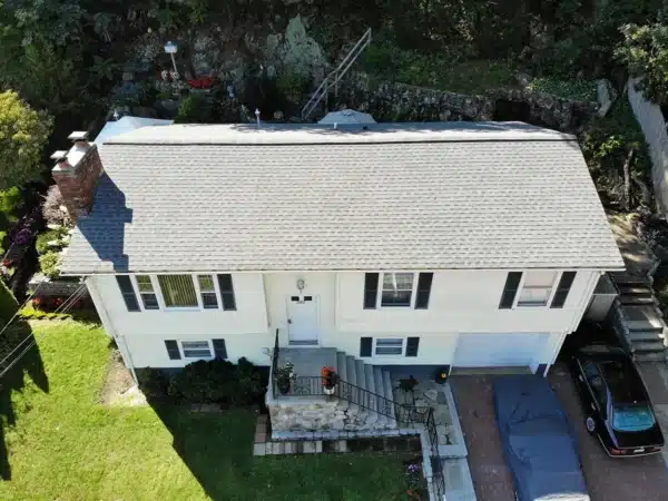 Aerial view of a white two-story house with a gray roof ideal for summer inspection. The dark-shuttered home features a stone staircase leading to the front door. A driveway hosts two cars, one covered, surrounded by lush lawns and trees, creating an inviting ambiance.