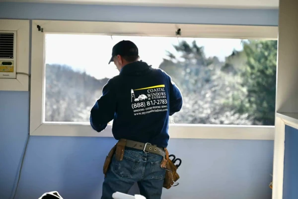 A man in a blue hoodie, cap, and tool belt expertly handles window installation in MA. The hoodie displays a Coastal Windows logo and phone number. He stands in a sunlit room with blue walls, facing a large open window framing the trees outside.