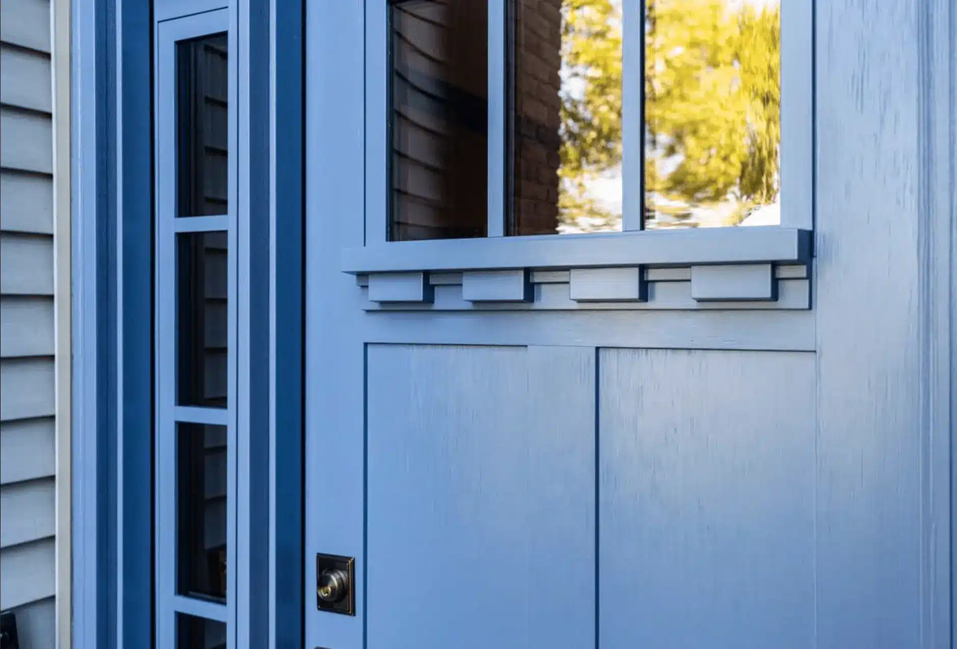 Close-up of a blue front door with glass panels and decorative trim. The panels reflect nearby trees, suggesting a sunny day. A brass doorknob is visible on the left. The door is framed by light gray siding.