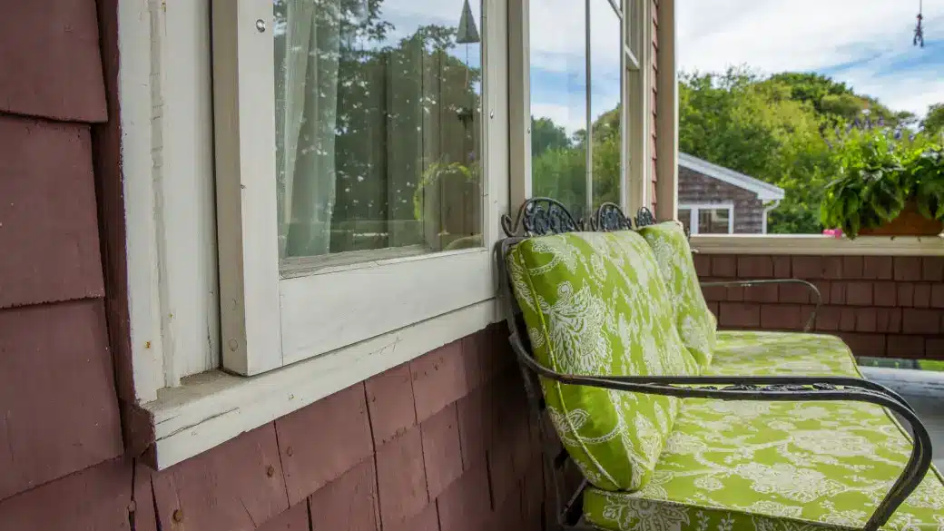A close-up view of a porch with two green cushioned chairs with patterned fabric, positioned against a house with reddish-brown shingles and white window trim. The window seal stands out as a potted plant and trees are visible in the background.