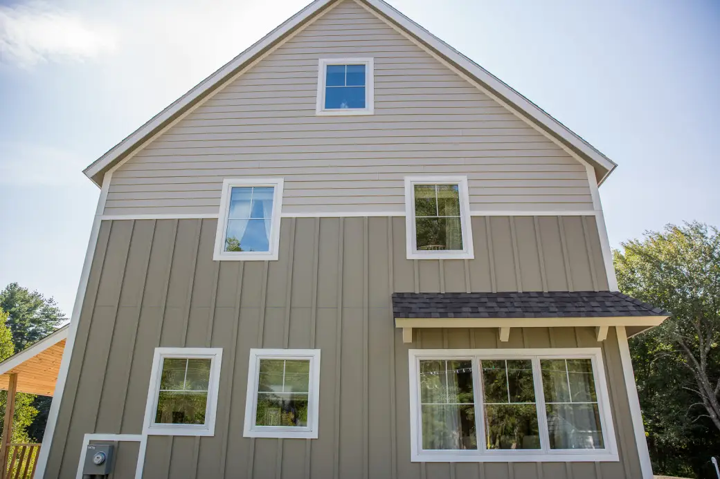 The Design Home Project showcases a two-story house with beige and light brown siding, accented by multiple white-framed windows from Coastal Windows & Exteriors. A small overhang graces the large ground-floor window, framed by trees and a clear blue sky—a vision worthy of Boston magazines pages.