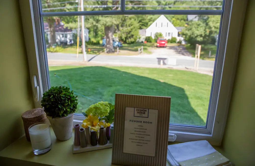 View from a window showing a suburban street with a red car and houses. The foreground boasts an eco-friendly vibe on the windowsill, featuring decorative items, including a leafy plant, a candle, flowers, and a framed sign labeled POWDER ROOM. Its all about going green!