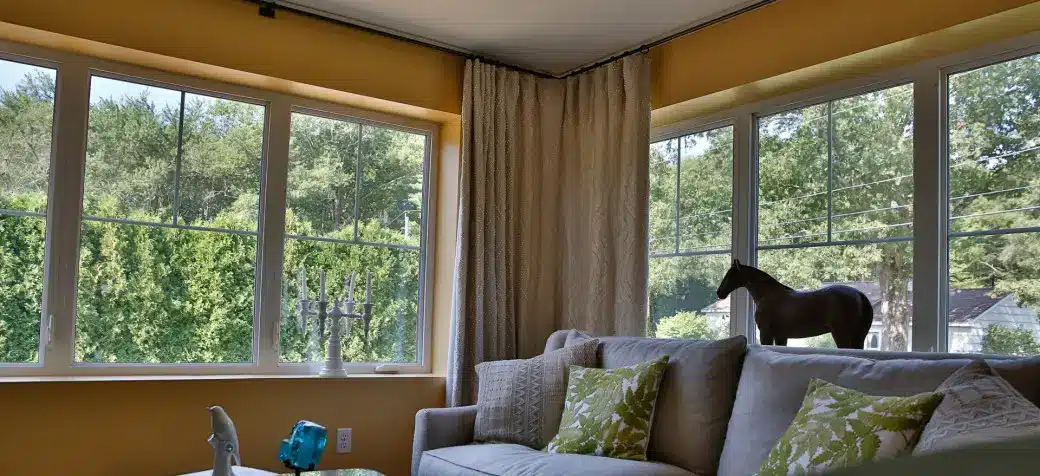 Bright living room with large corner windows featuring stylish shades, yellow walls, a light gray sofa with green patterned pillows, a table, and a decorative horse figurine on the window ledge; trees visible outside.