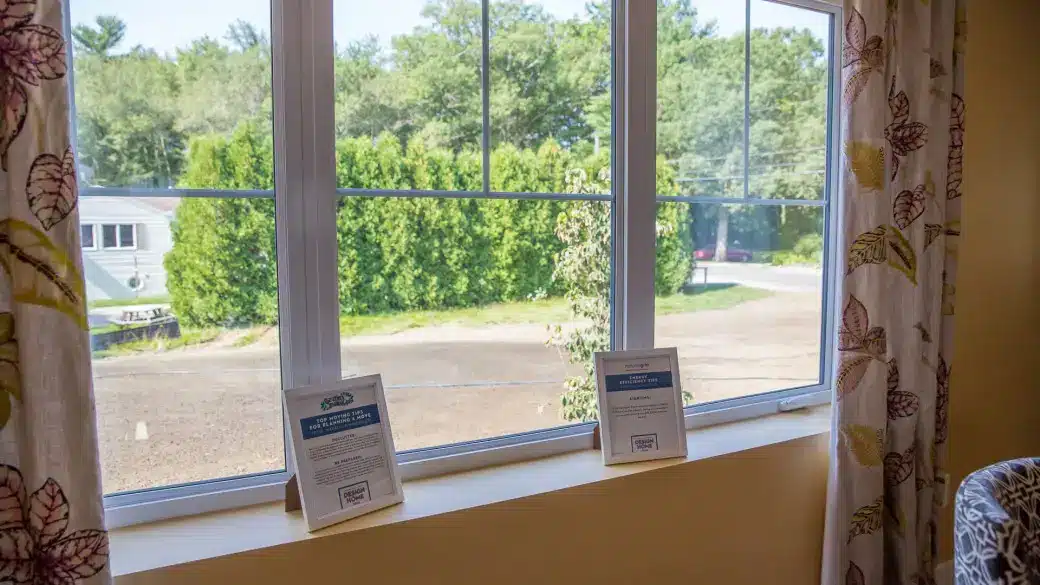 A large window with floral curtains overlooks a sparsely wooded area, reminiscent of an expensive project completed at Westford School. On the windowsill are two small framed signs. Outside, a car and trees are visible under a clear blue sky.